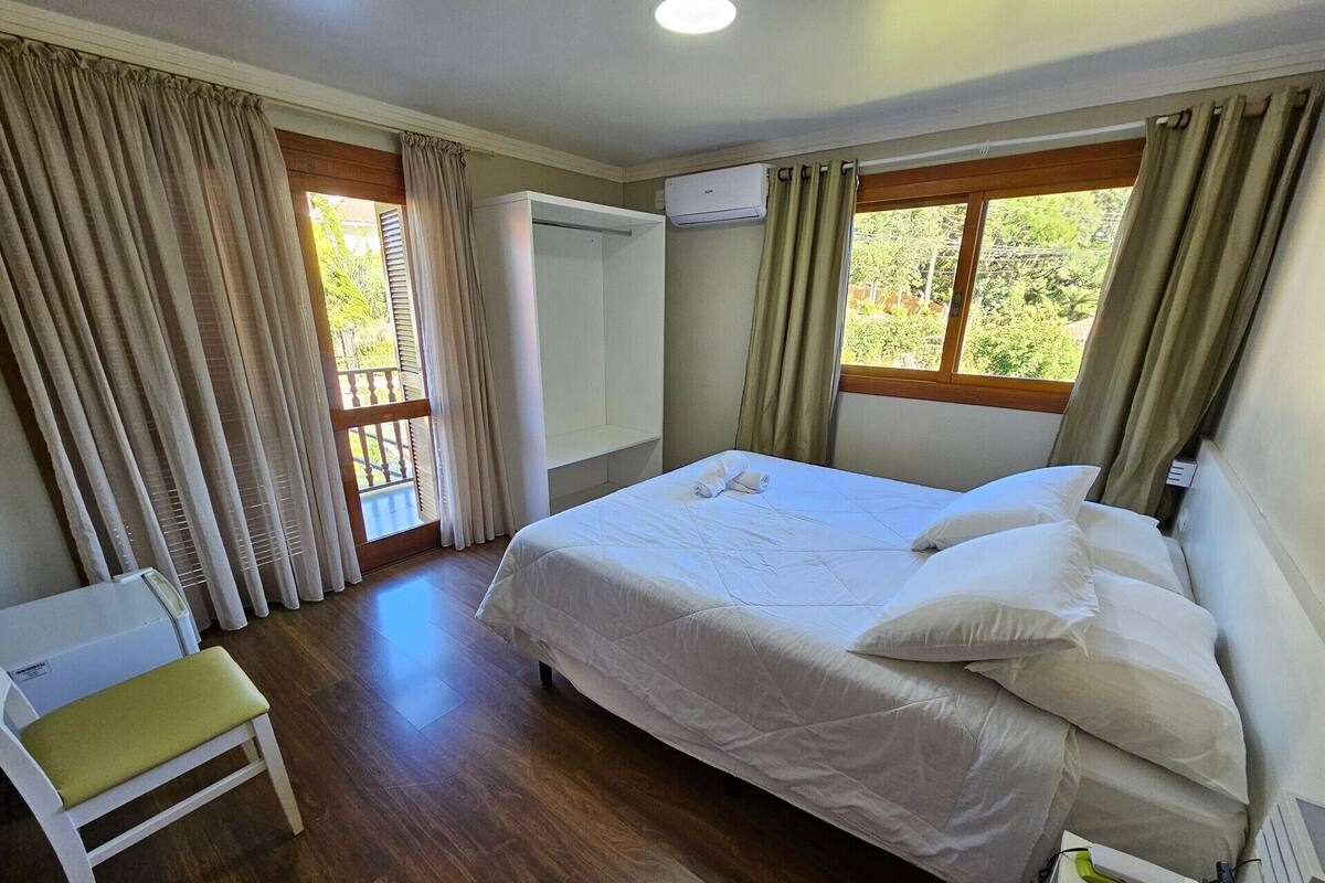 Un dormitorio con una cama, una silla, un ventanal con cortinas y un equipo de aire acondicionado.