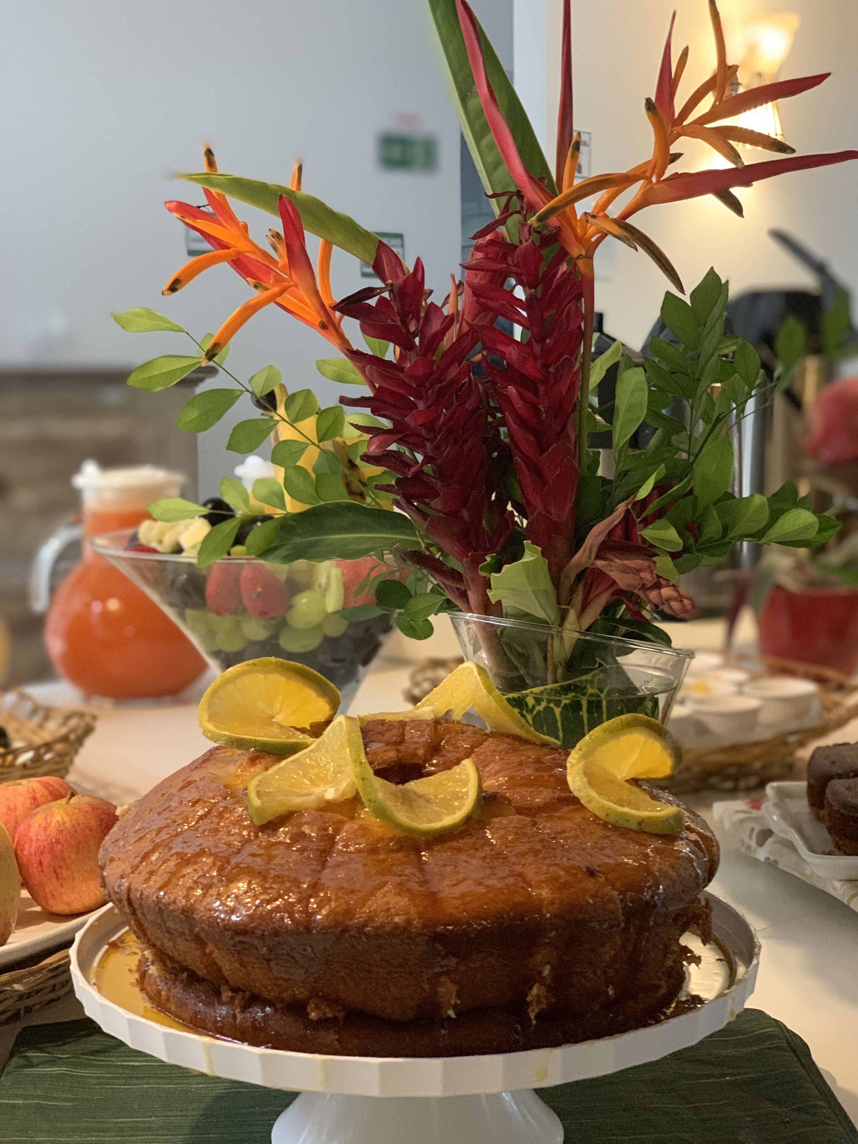 A dessert cake with lime slices on a white stand, surrounded by a variety of fruits and a decorative plant arrangement.