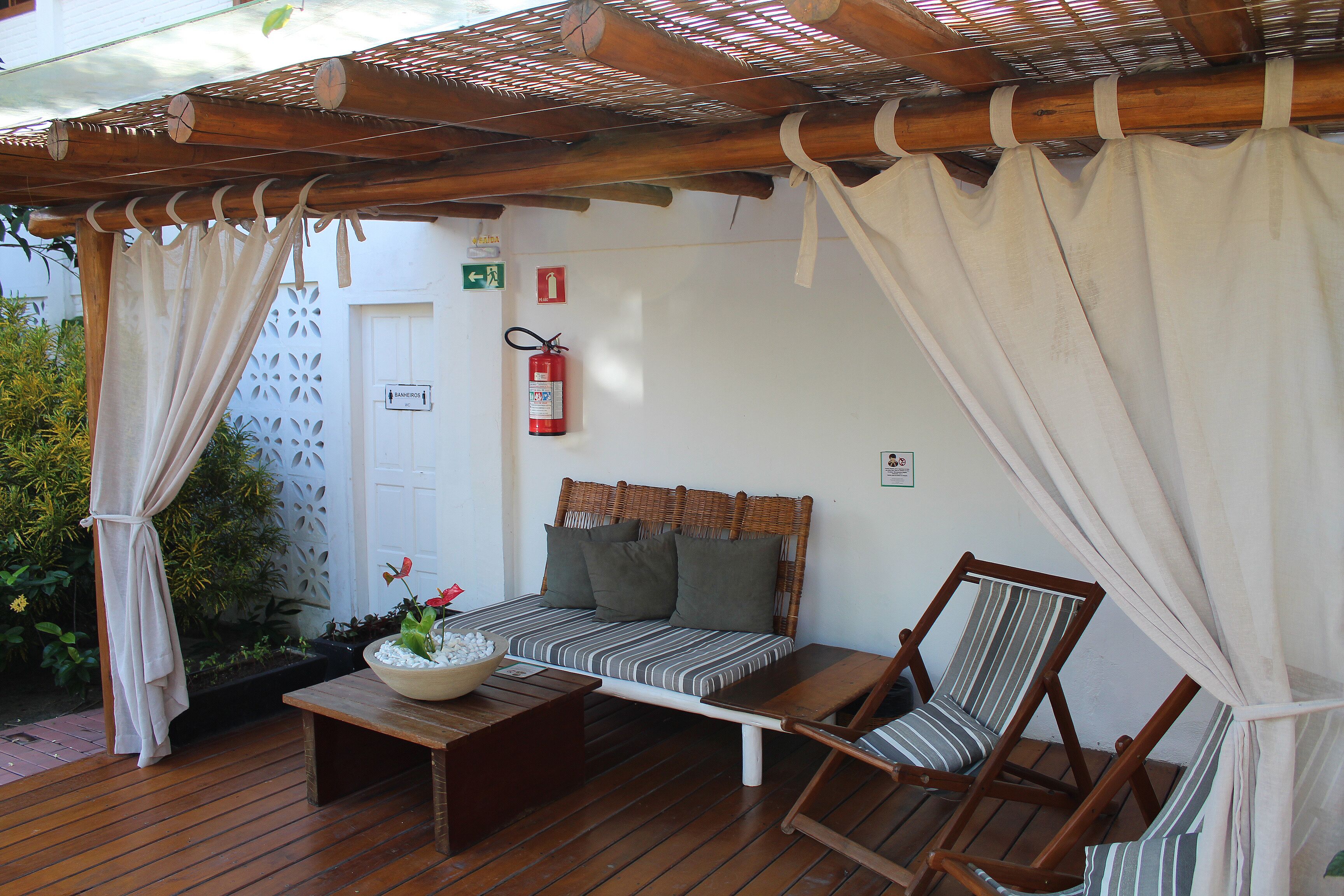 A covered outdoor seating area with a wooden bench, a cushioned bench, and a table with a bowl of flowers.
