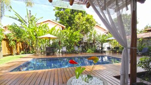 Une terrasse en bois avec une piscine, entourée d’une végétation luxuriante et dotée d’un espace salon abrité.