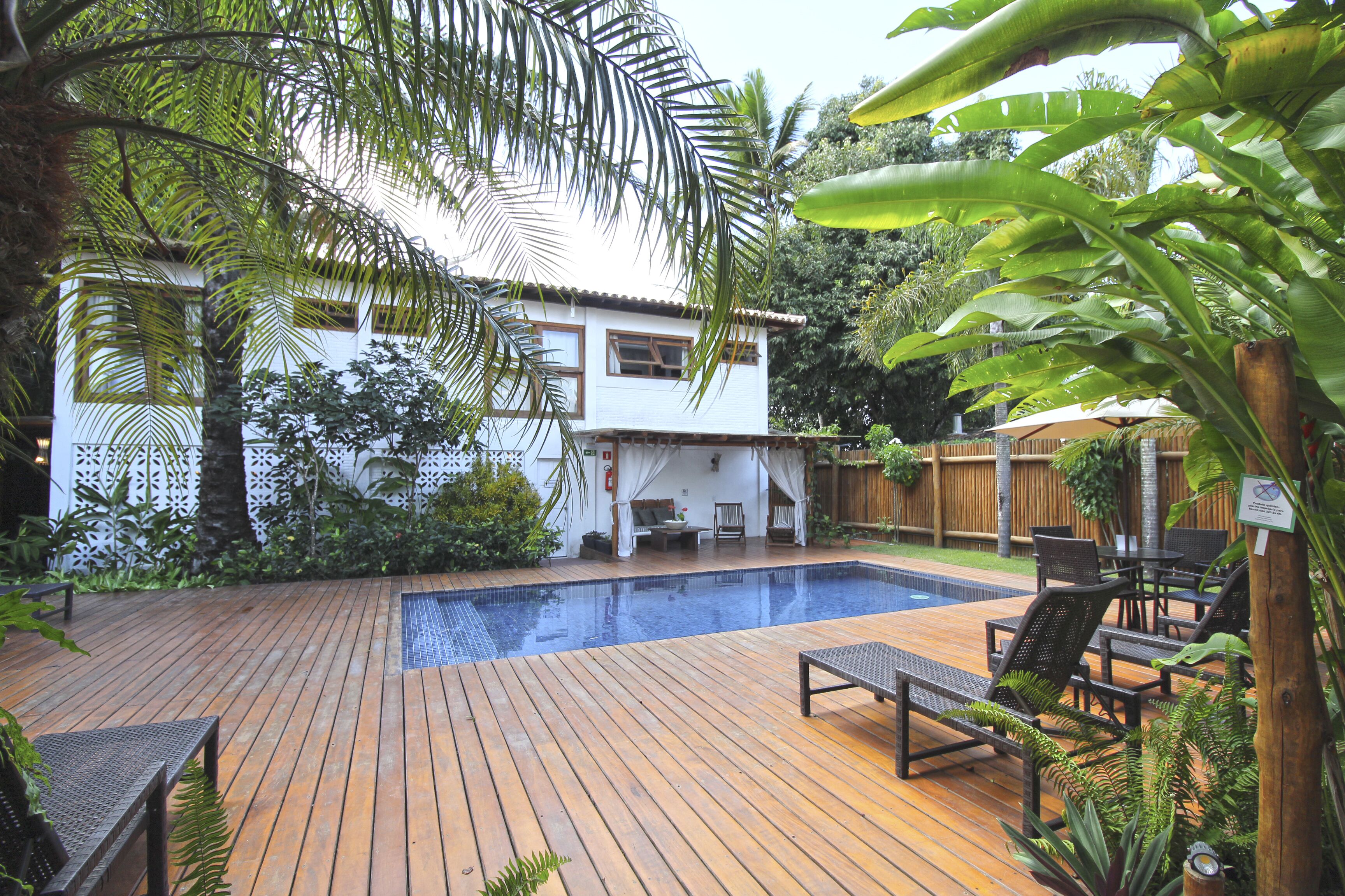 Um deck de madeira com piscina, cercado por vegetação exuberante e uma casa branca.