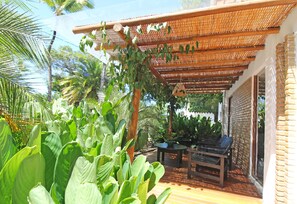 Un espace extérieur aménagé, abrité et doté d’une terrasse en bois, entouré d’une végétation luxuriante et d’un mur en briques.