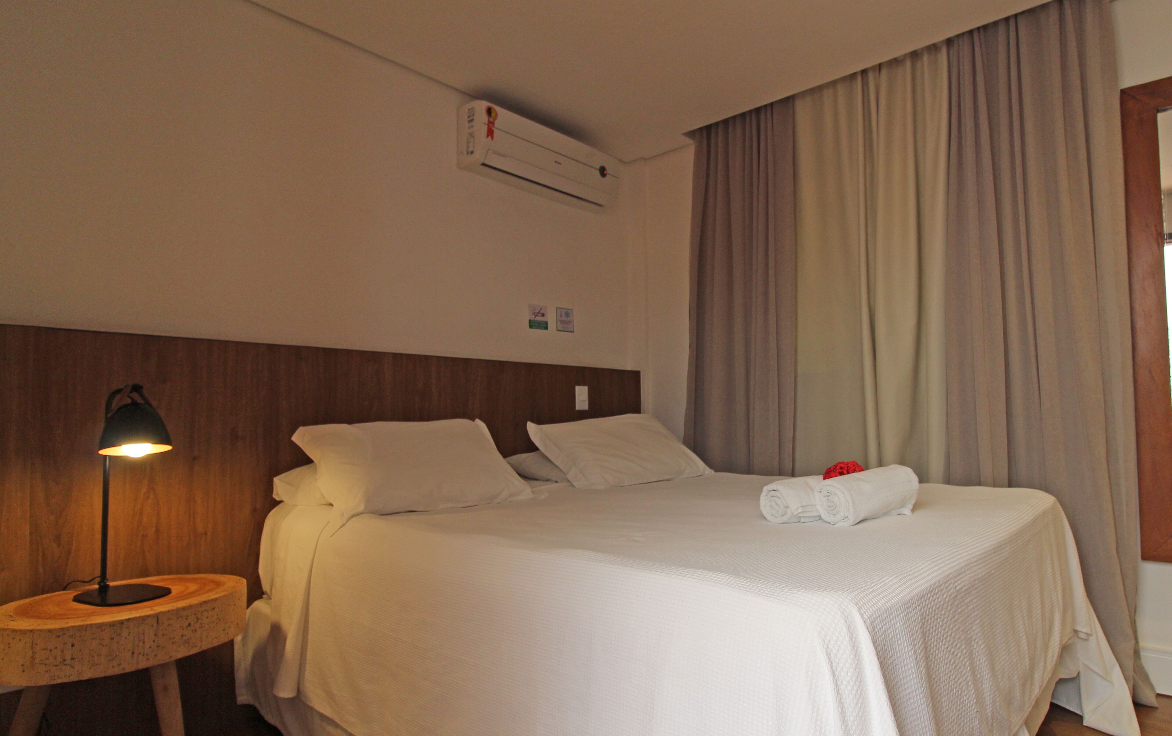 A hotel room with a neatly made bed, a round wooden side table with a lamp, and a large window with curtains.