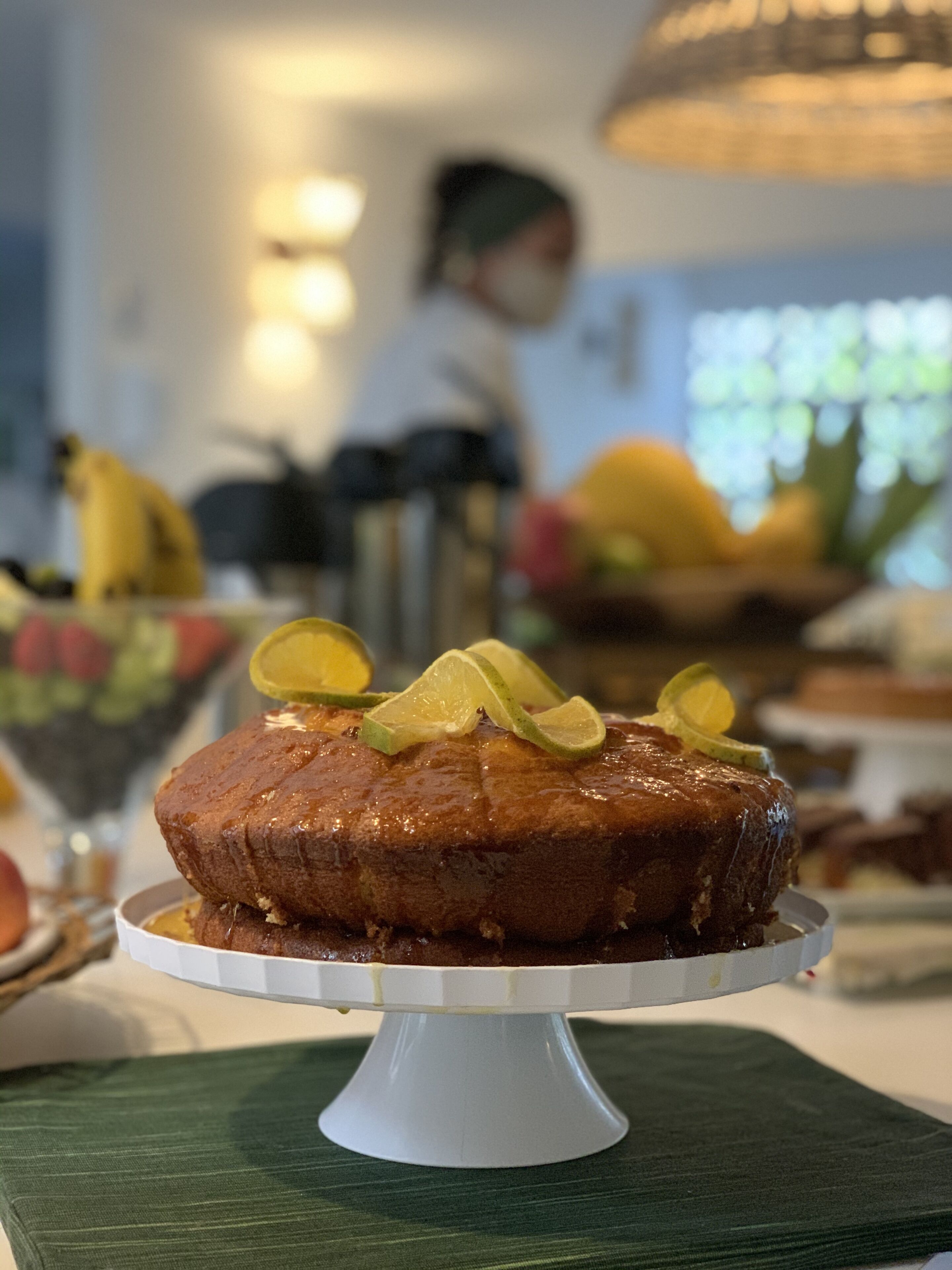 A cake on a white stand with lime slices on top, placed on a green cloth.