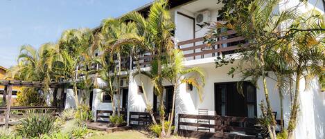 A two-story building with balconies, surrounded by palm trees and a garden area with wooden benches.