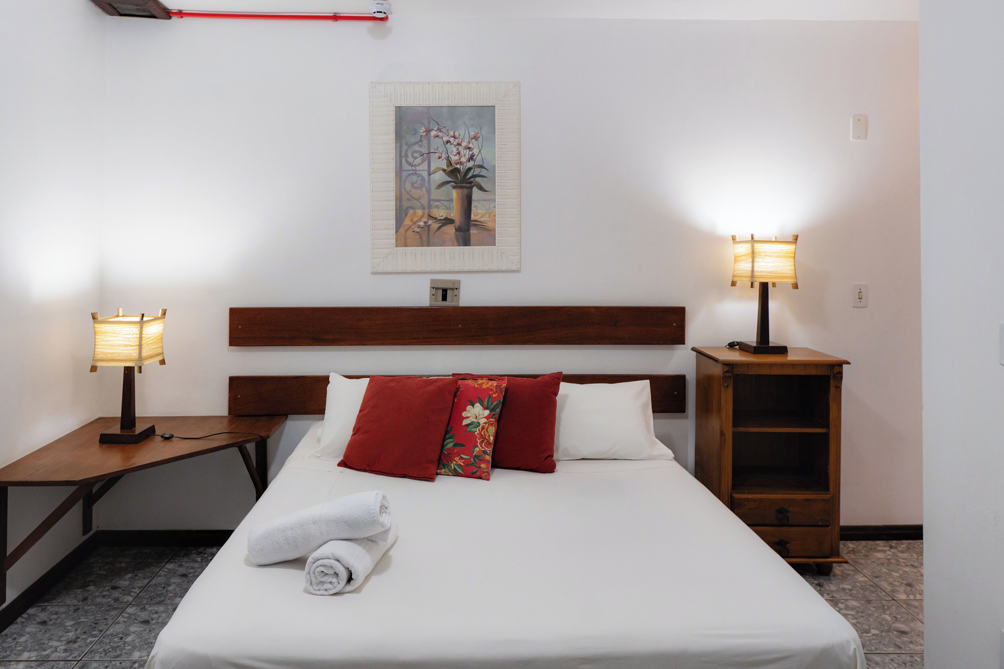 A hotel room with a neatly made bed, two bedside lamps, a wooden nightstand, a small wooden cabinet, and a painting on the wall.