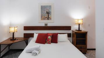 A hotel room with a neatly made bed, two bedside lamps, a wooden nightstand, a small wooden cabinet, and a painting on the wall.