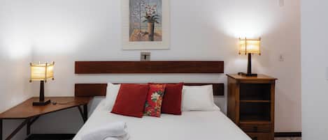 A hotel room with a neatly made bed, two bedside lamps, a wooden nightstand, a small wooden cabinet, and a painting on the wall.