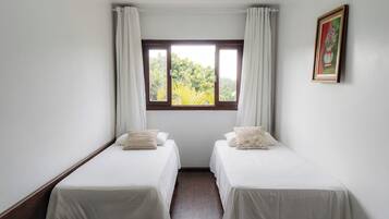A room with two single beds, white curtains, a window with a view of greenery, and a framed picture on the wall.