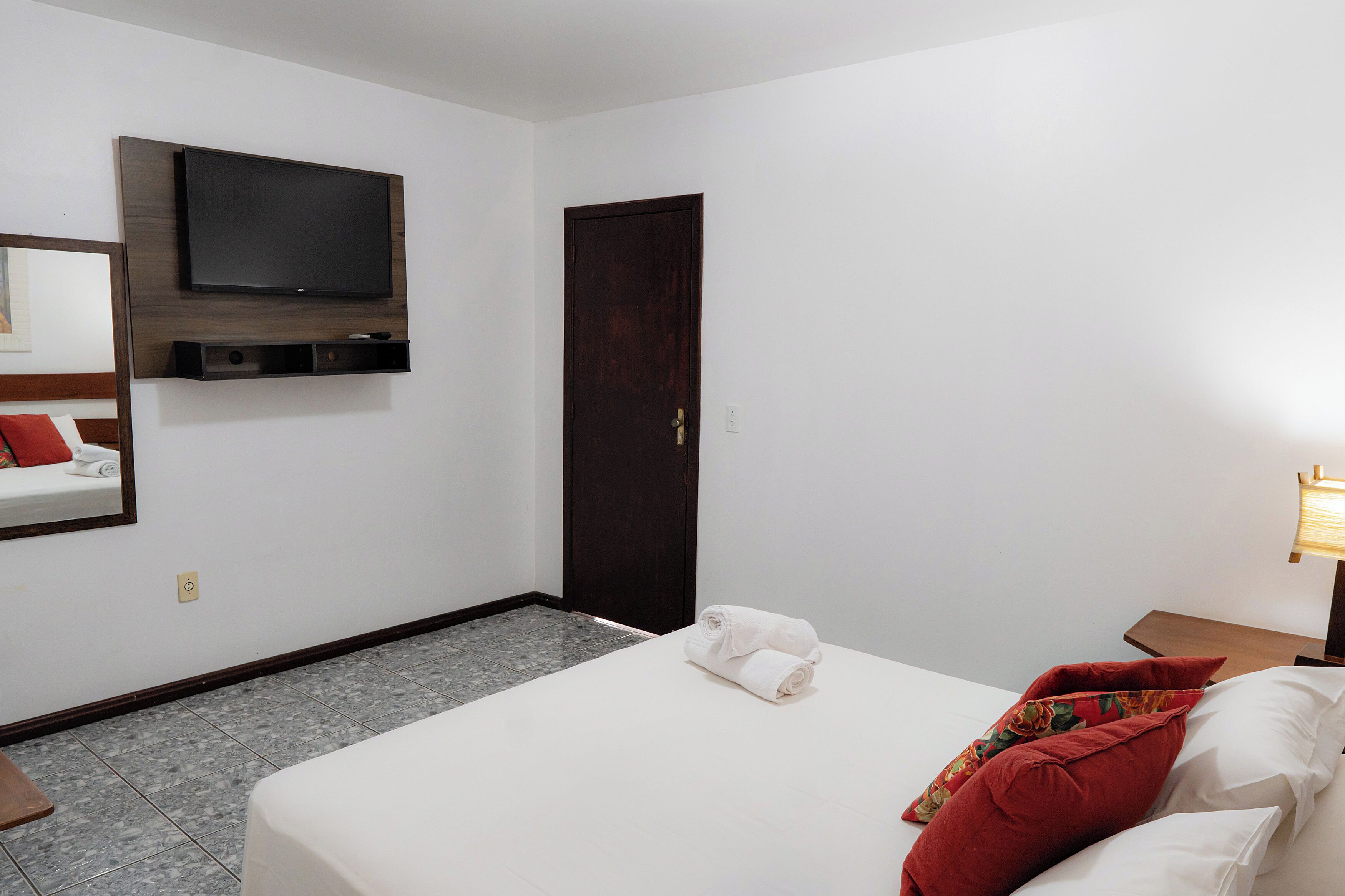Una habitación de hotel con una cama, televisión montada en la pared, una puerta y una mesita de noche con lámpara.