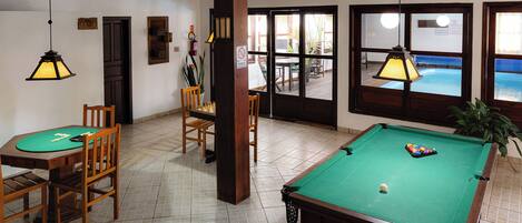 A pool table with a green felt surface, wooden chairs, and a small table with a green felt tablecloth.