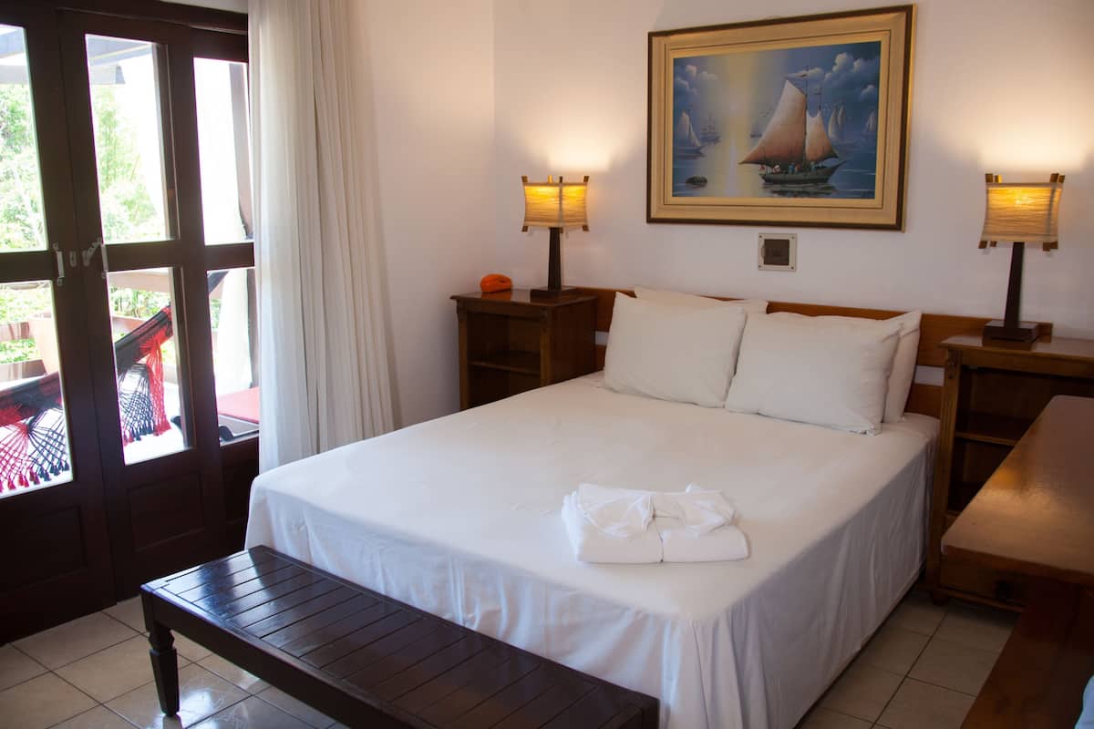 A hotel room with a bed, two bedside tables with lamps, a bench, a painting on the wall, and a view through a door.