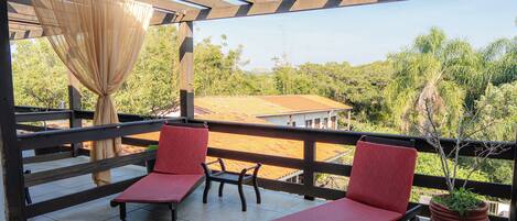 A covered outdoor seating area with two red lounge chairs and a small table, overlooking a landscaped garden and a building with a red roof.