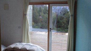 Un dormitori amb un llit, una porta corredissa de vidre que dona a una terrassa exterior, i vista a arbres i una casa al fons.