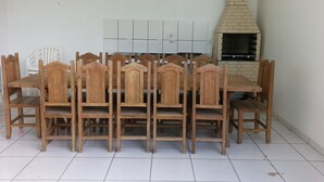 A dining area with a wooden table and chairs, a white chair, and a brick oven in the background. - Residencial Floripamar 2 (Florianópolis)