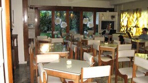 Restaurante com mesas e cadeiras de madeira, um ventilador de teto e vista para o exterior através de amplas janelas.