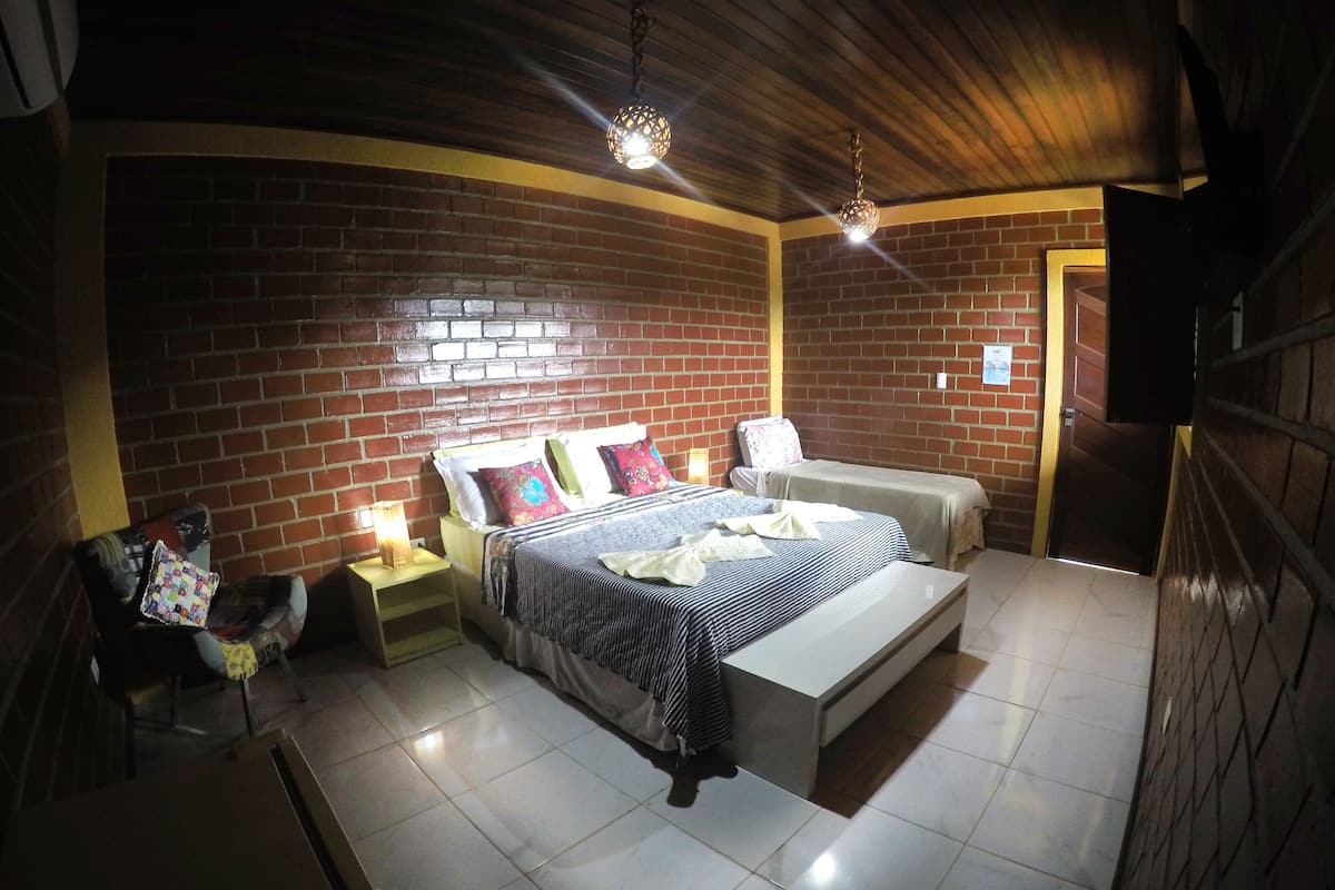 Quarto com parede de tijolos, cama com colcha listrada, um banquinho e mesa de cabeceira com abajur.