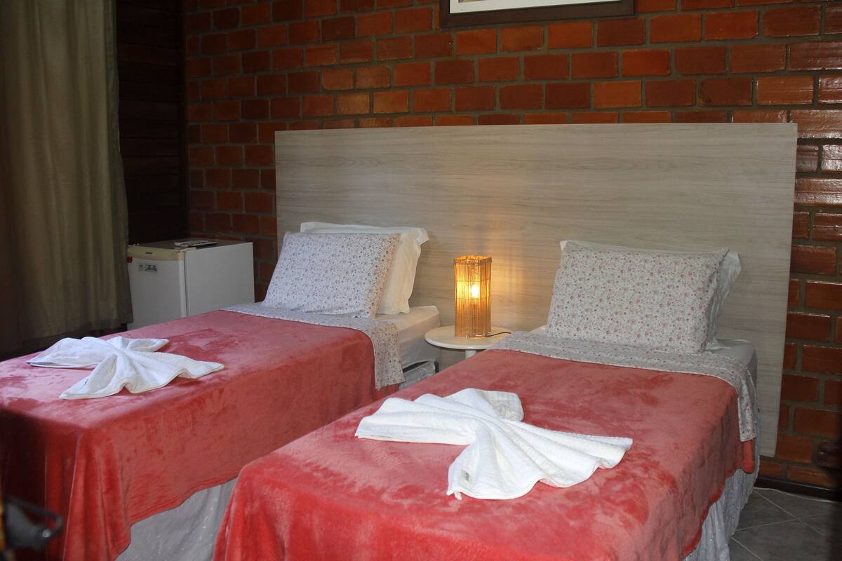 Quarto de hotel com duas camas, cada uma com colcha vermelha e toalhas brancas. Há uma pequena mesa entre as camas com um abajur. O quarto tem uma parede de tijolos e uma cabeceira de madeira.