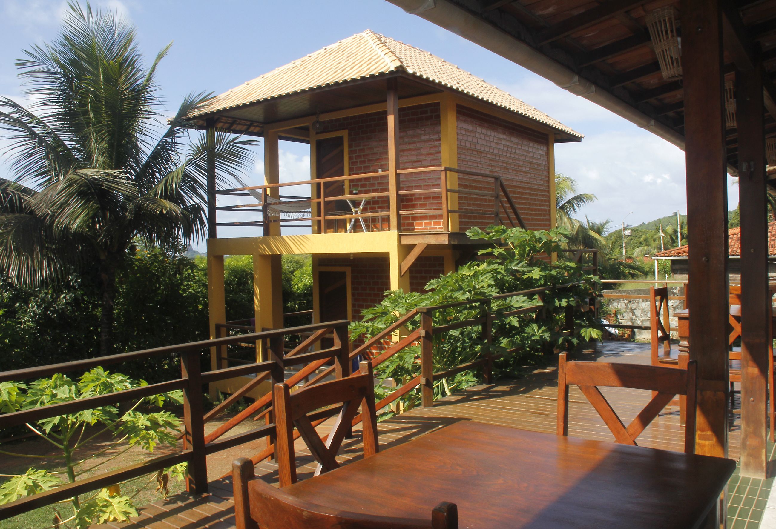 A wooden deck with a table and chairs, a small wooden structure with a tiled roof, and lush greenery in the background.