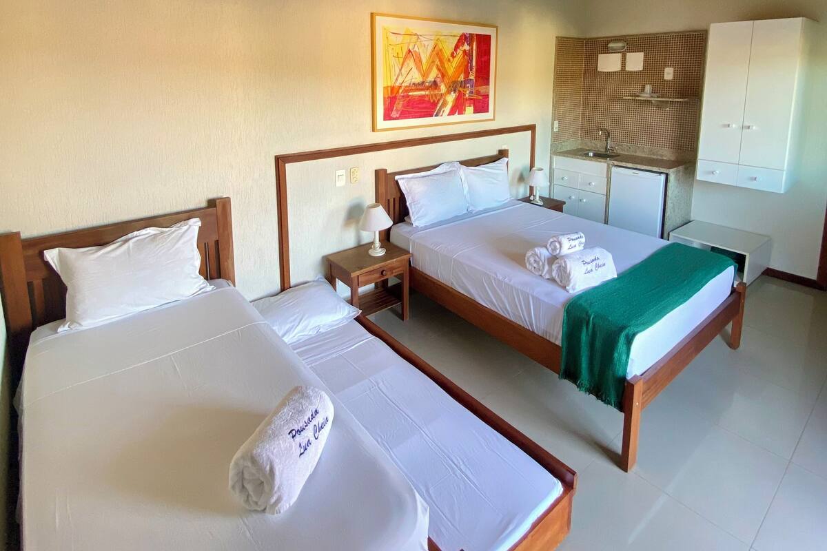 A hotel room with two single beds, a wooden ceiling, a painting on the wall, a kitchenette, and a white wardrobe.