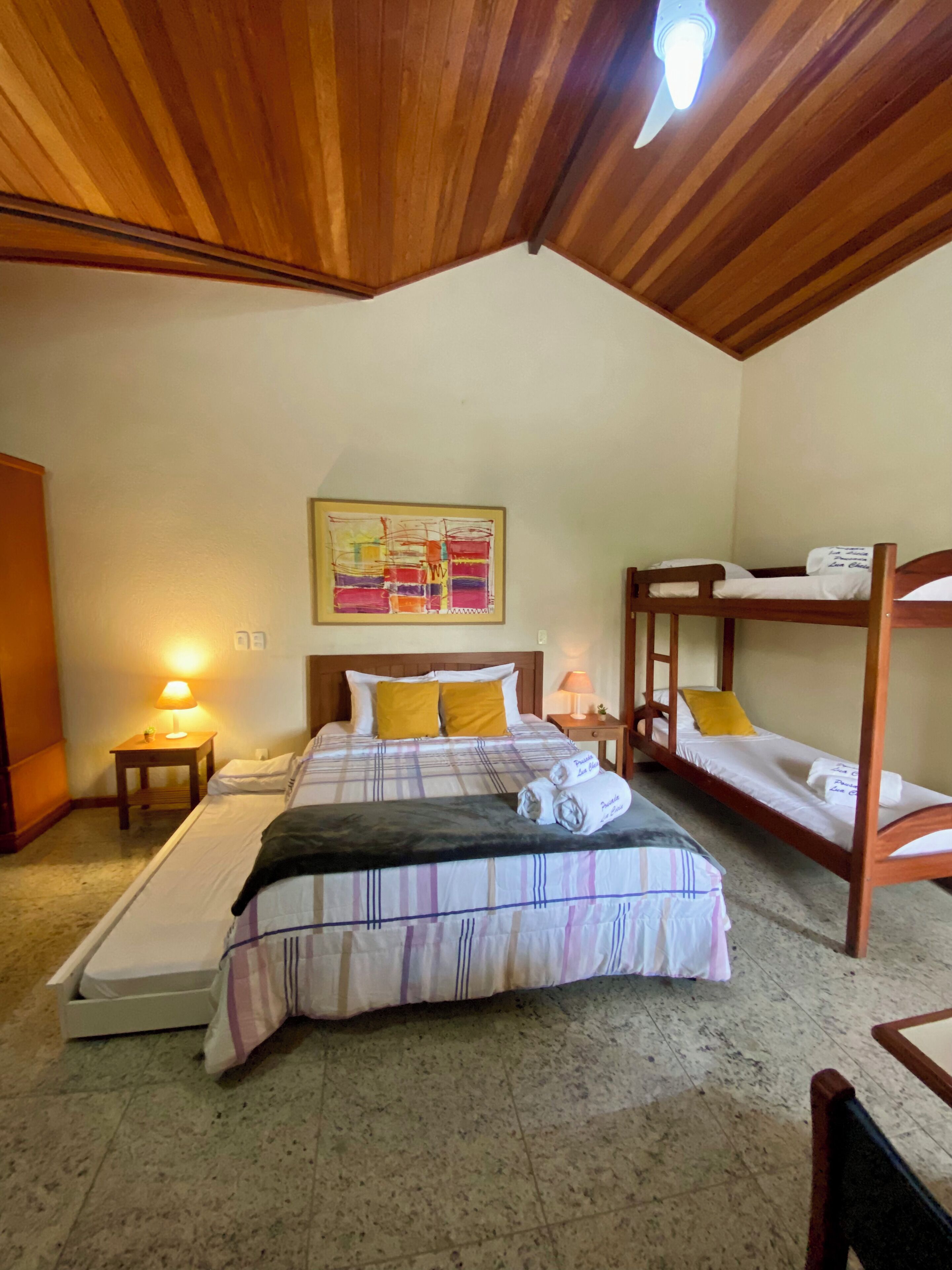 Una habitació d'hotel amb un llit gran, dues taules de nit, un llit superposat de fusta, un ventilador de sostre i un quadre a la paret.