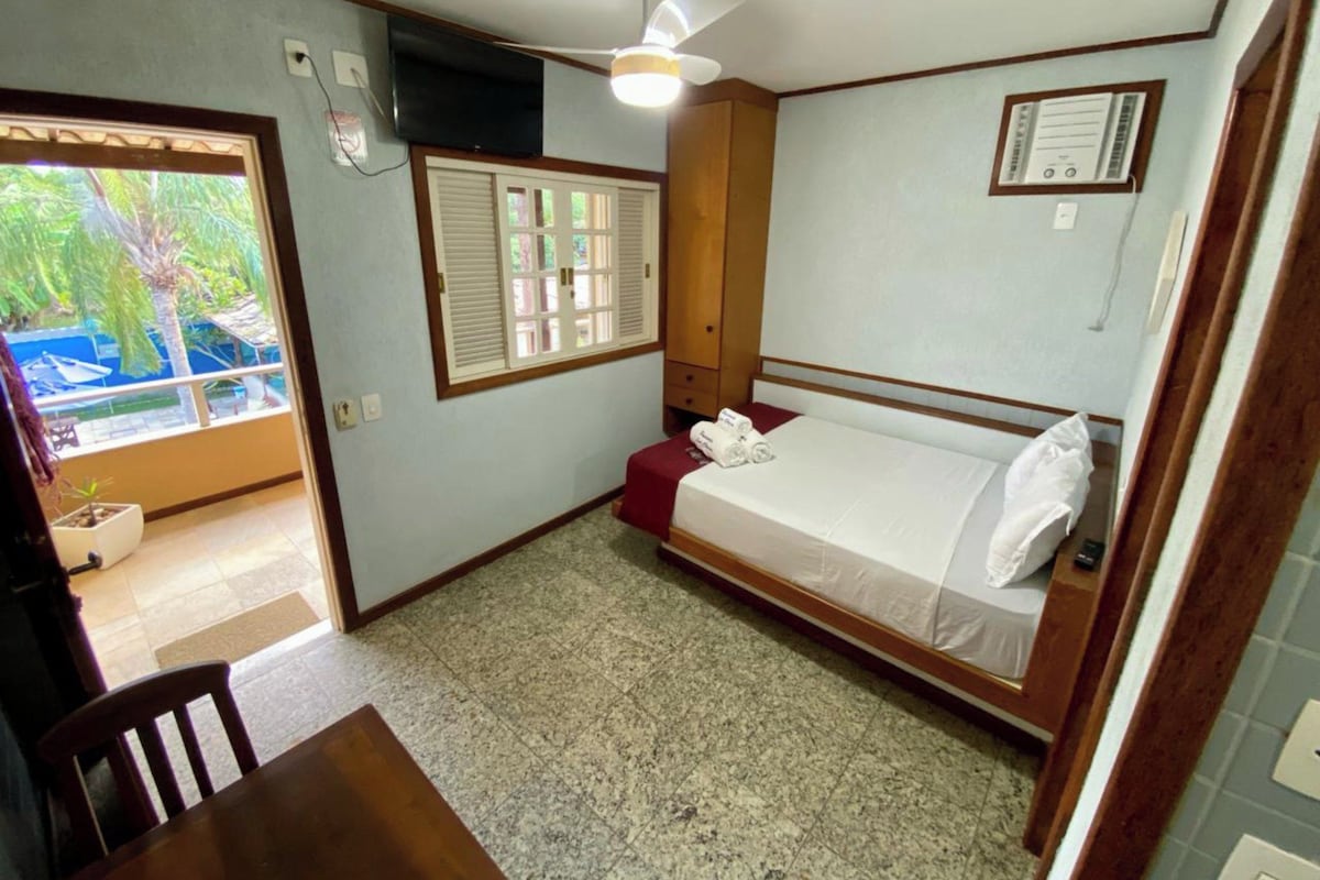 Una habitación pequeña con una cama, un ventilador de techo, una ventana con persianas, un televisor y una mesita.