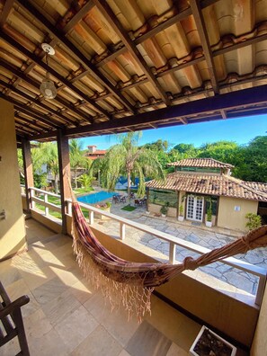 A balcony with a hammock, a pool, and a view of a house with a tiled roof. - Pousada Lua Cheia (Búzios)
