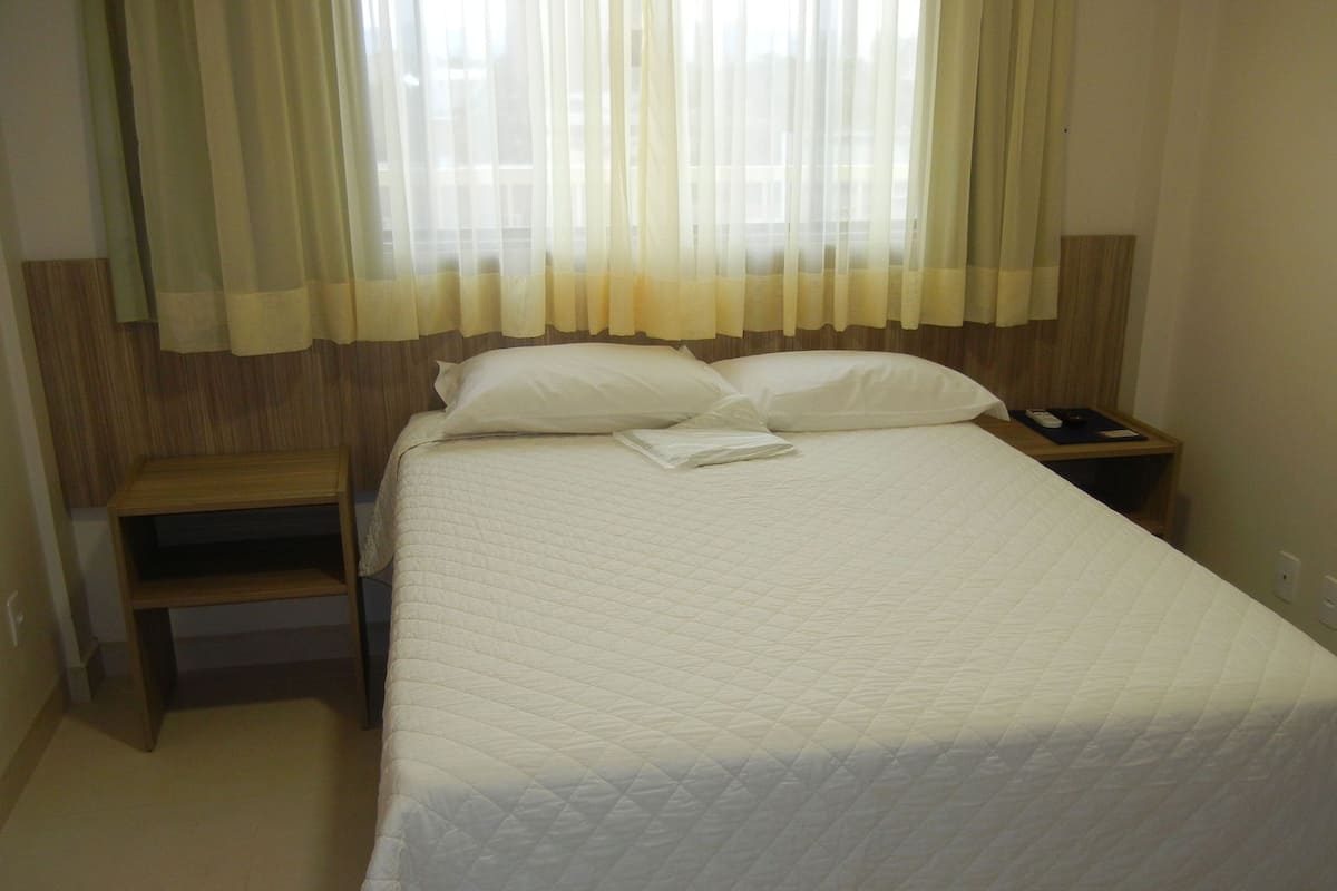 Un lit bien fait, recouvert d’une couette blanche matelassée, une table de chevet en bois avec une télécommande, et une chaise en bois placée à côté du lit.