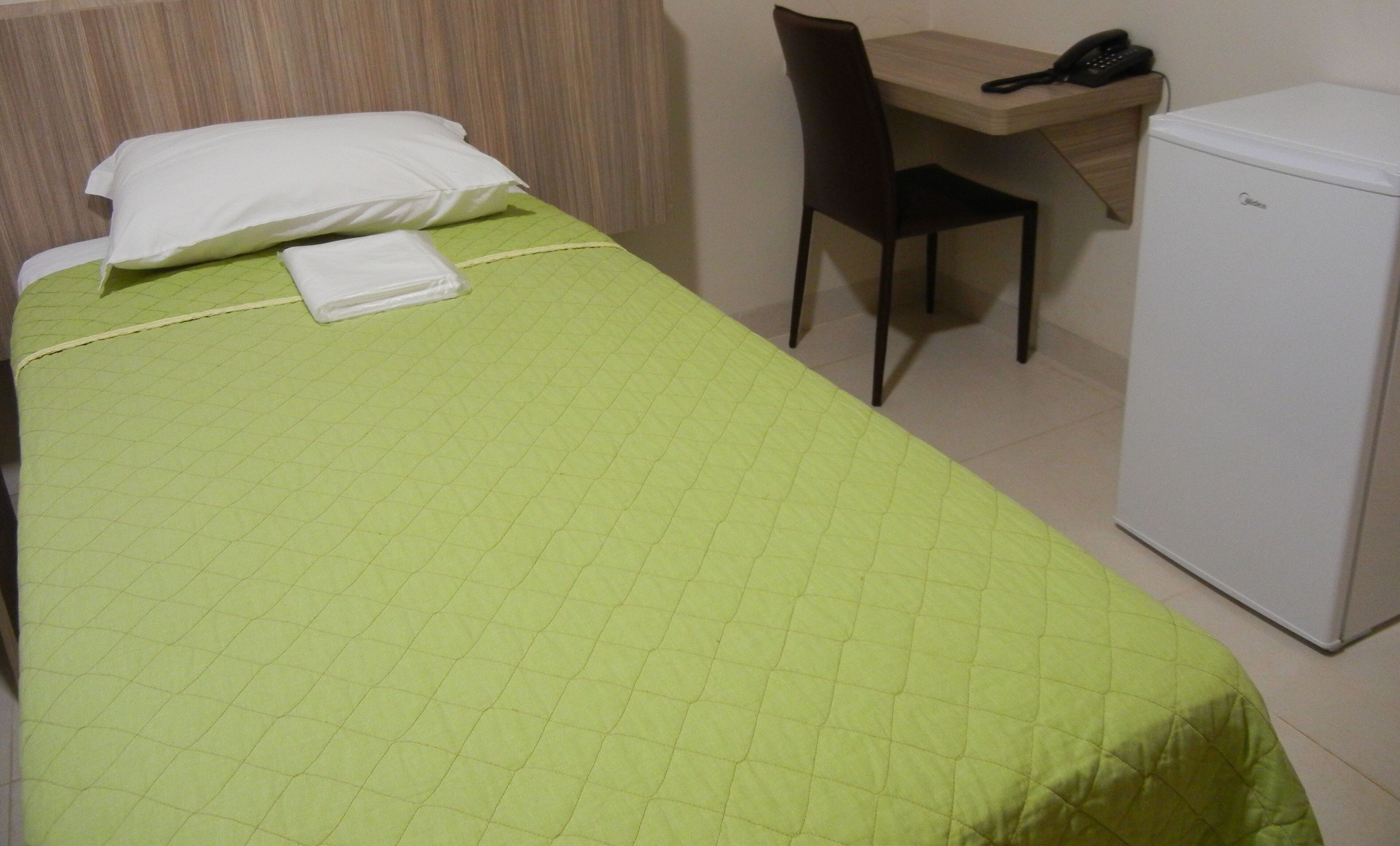 A single bed with a green quilt, a white pillow, and a folded towel. There is a desk with a chair and a phone on it. A small white refrigerator is next to the desk.