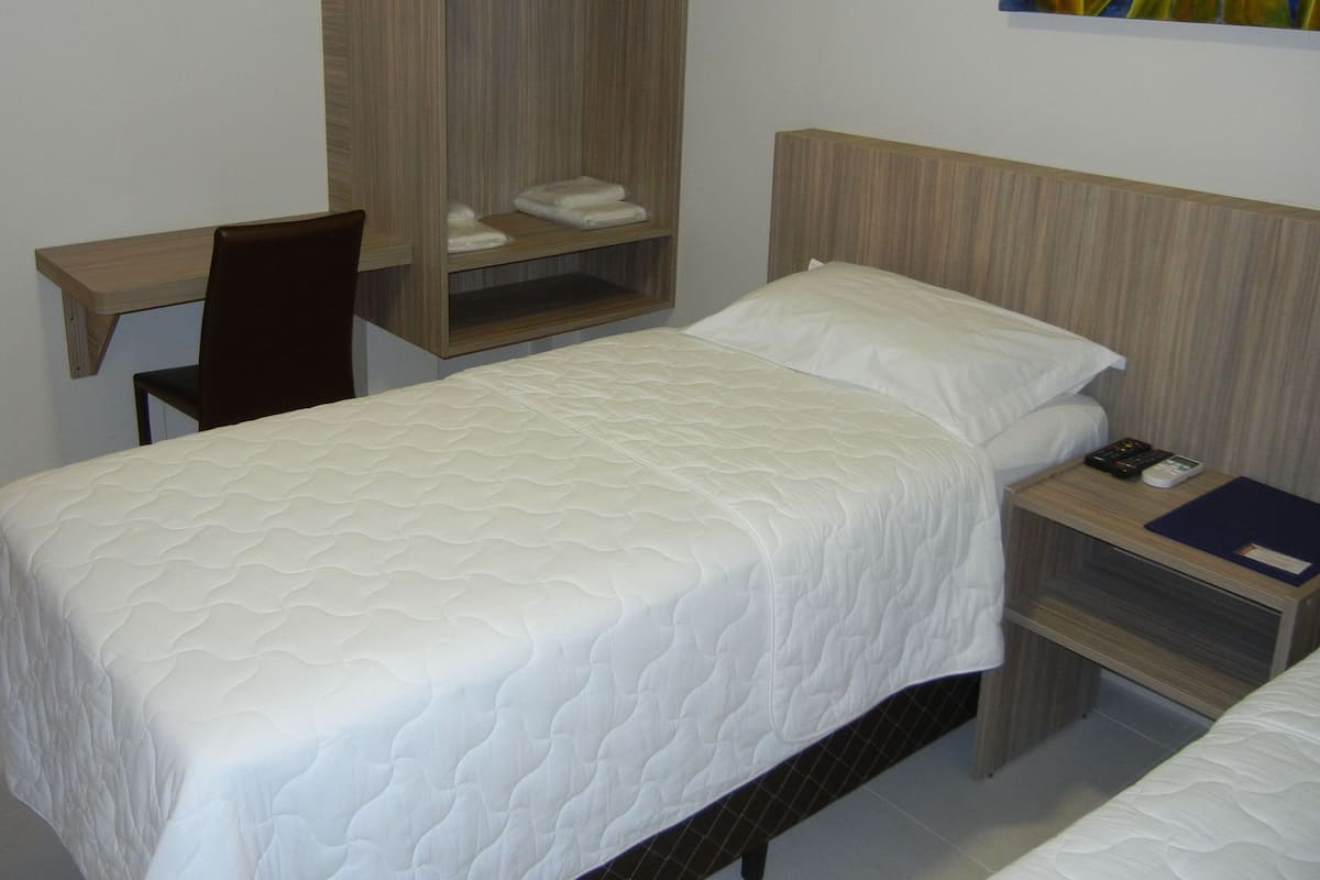Un lit simple avec une couette blanche matelassée, une table de chevet en bois avec une télécommande et un livre, et une armoire en bois contenant des serviettes pliées.