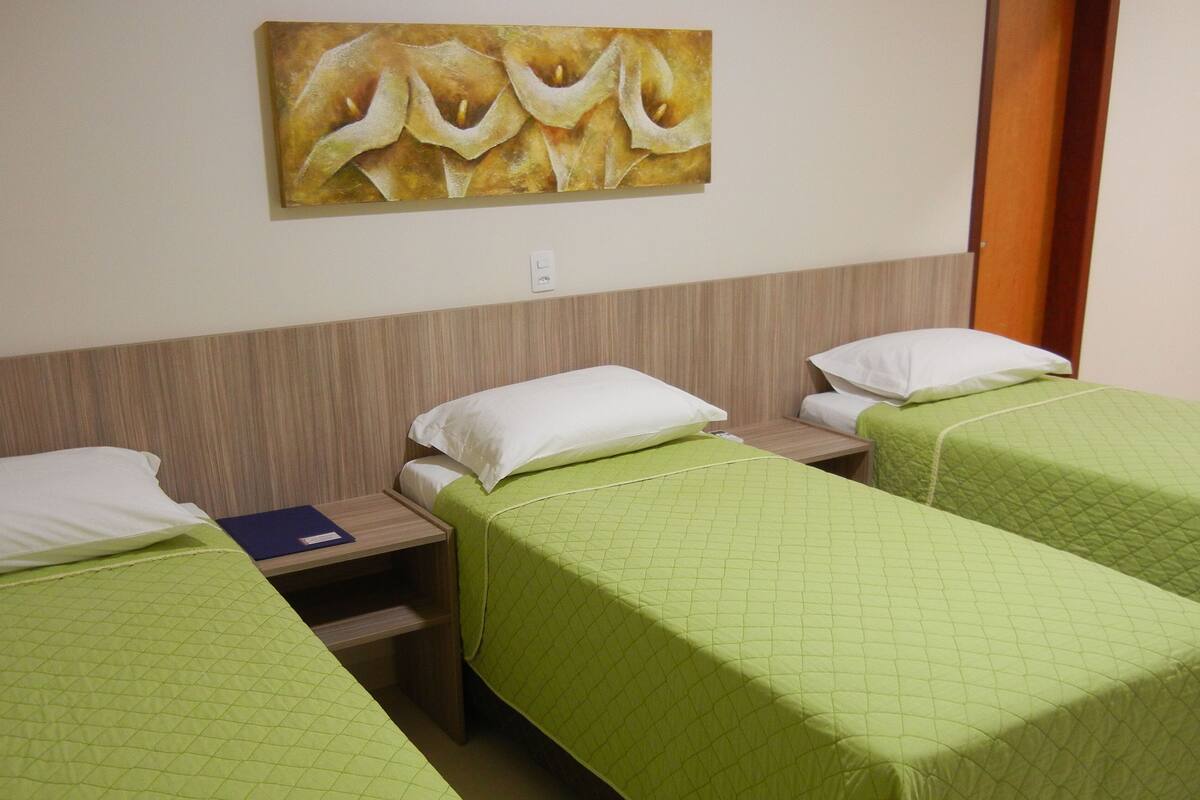 Une chambre d’hôtel avec deux lits simples, chacun recouvert d’une courtepointe verte matelassée et agrémenté d’oreillers blancs. On y trouve une tête de lit en bois, une petite table avec un dossier bleu et un tableau accroché au mur.