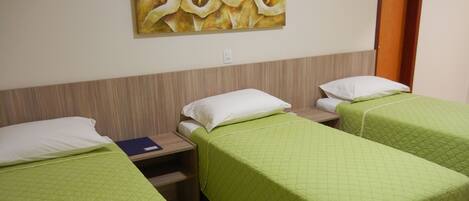 Habitación de hotel con dos camas individuales, cada una con una colcha verde acolchada y almohadas blancas. Se observa un cabecero de madera, una pequeña mesa con una carpeta azul y un cuadro en la pared.