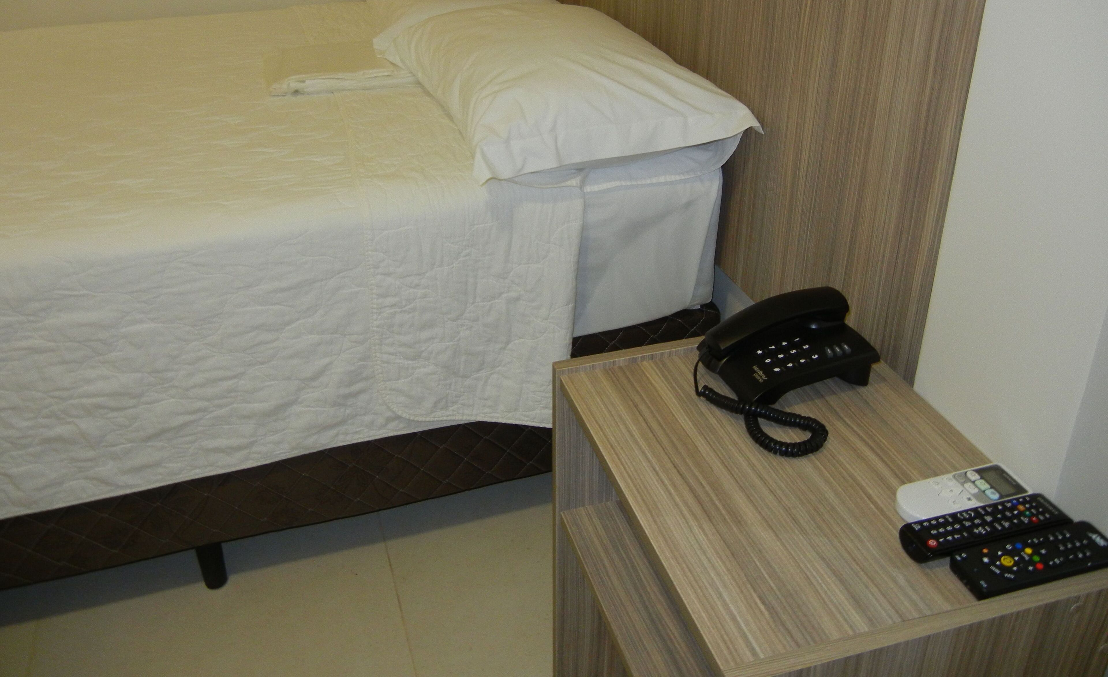 A hotel room with a bed, a bedside table with a telephone and remote controls, and a wall-mounted light switch.