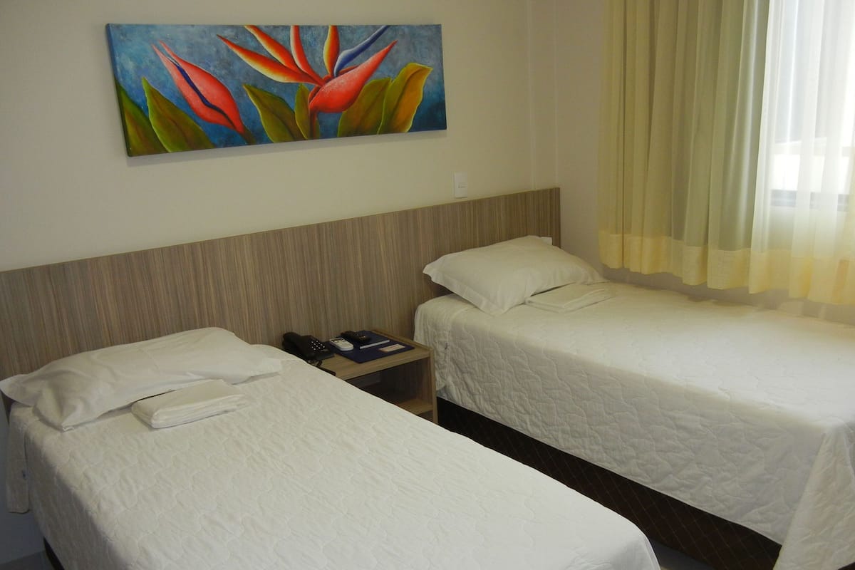 Une chambre d’hôtel avec deux lits simples, une table de chevet avec un téléphone et une télécommande, et un tableau représentant des fleurs accroché au mur.