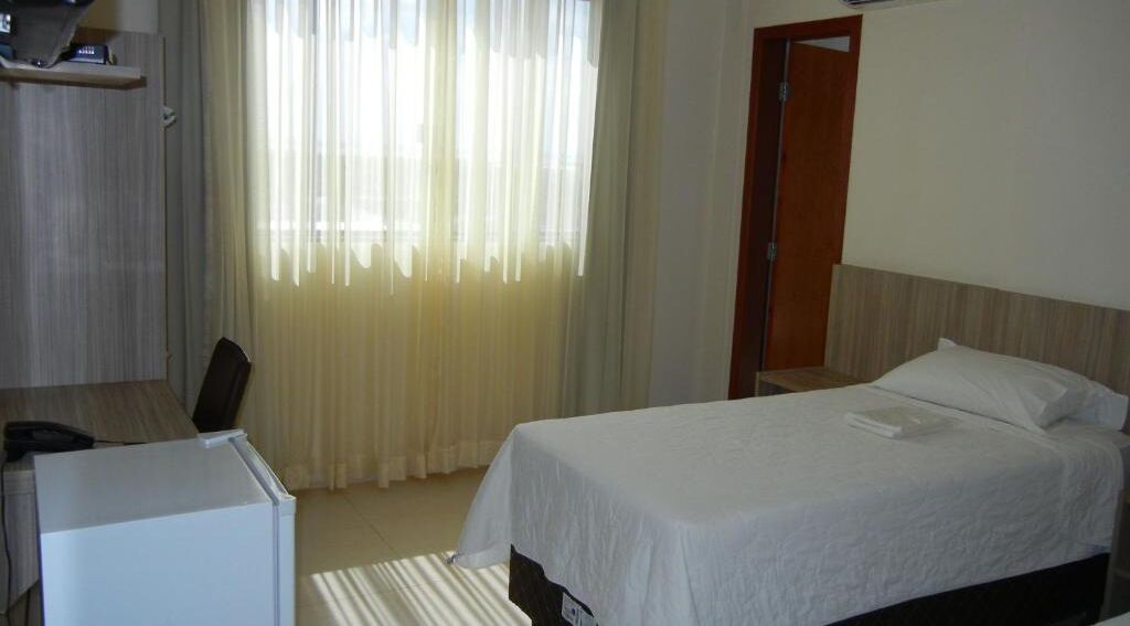 A hotel room with a bed, a desk, and a chair. There is a window with curtains, a door, and a small table with a lamp.