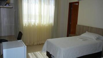 A hotel room with a bed, a desk, and a chair. There is a window with curtains, a door, and a small table with a lamp.