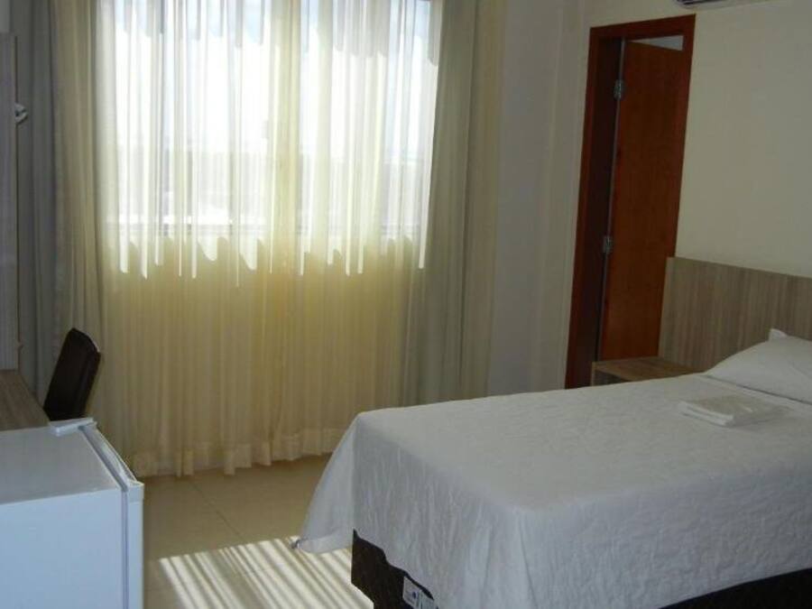 A hotel room with a bed, a desk, and a chair. There is a window with curtains, a door, and a small table with a lamp.