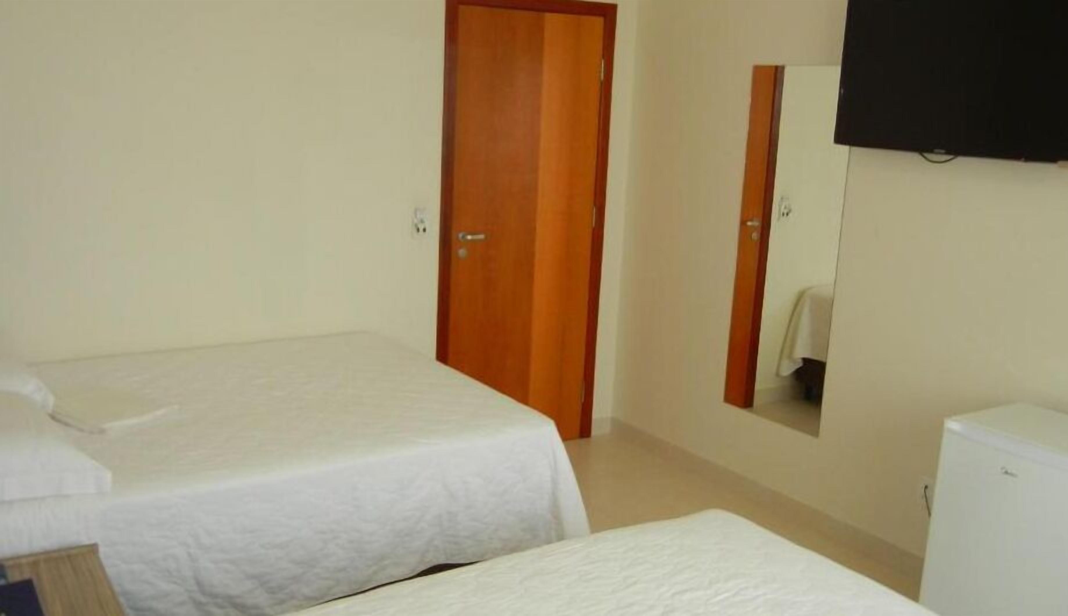 A room with two beds, a wooden door, a white refrigerator, and a mounted television.