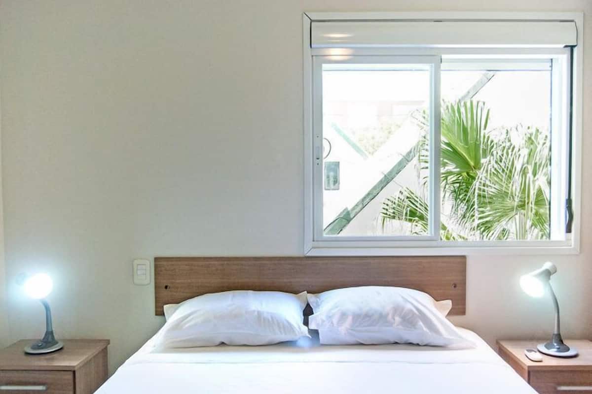 A bedroom with a bed, two bedside lamps, a window with a view of palm trees, and a wooden headboard.