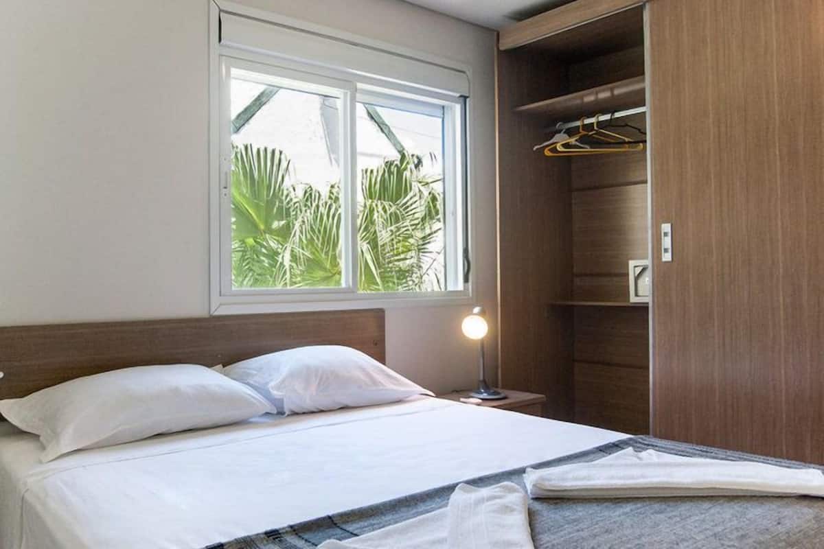A neatly made bed with white linens, a bedside lamp, and a window with a view of palm trees.