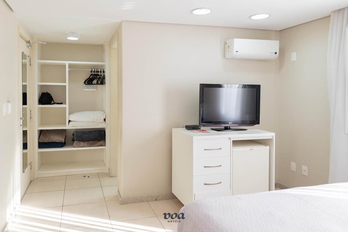 A hotel room with a bed, a TV on a white cabinet, a closet with hanging clothes, and a window with curtains.
