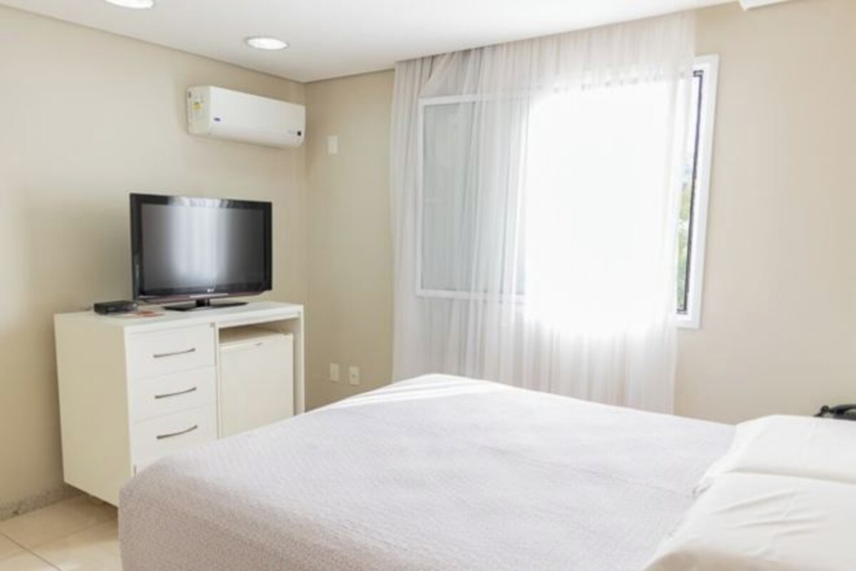 Una habitación de hotel con una cama, un televisor sobre un aparador blanco y una ventana con cortinas traslúcidas.