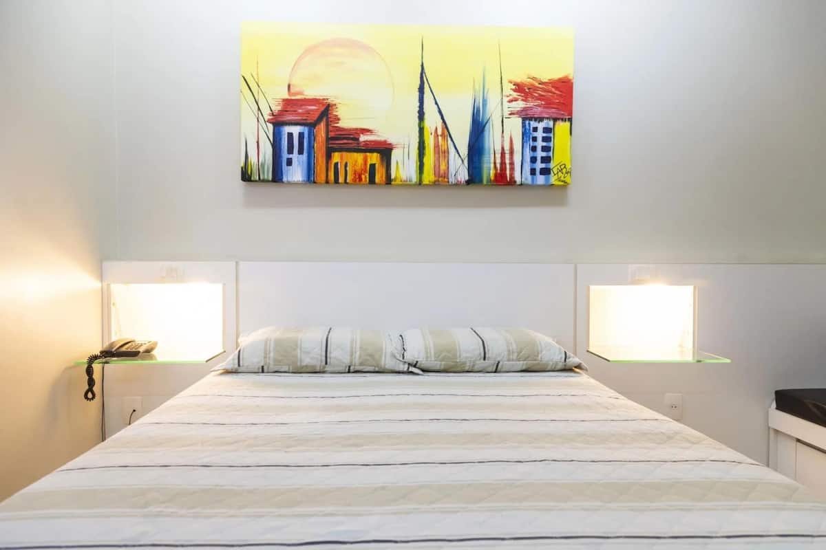A hotel room with a bed, bedside lamps, a painting on the wall, and a window.