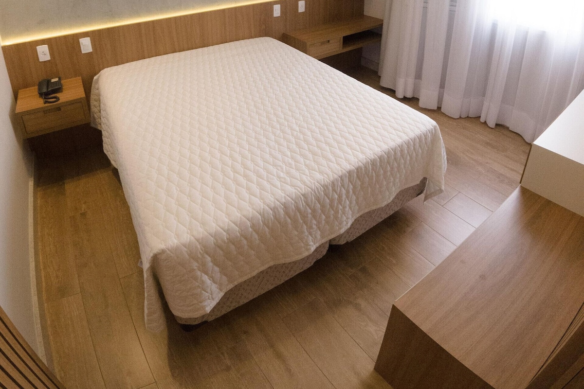 A modern hotel room with a large bed, wooden headboard, built-in shelving, and a large window with curtains.