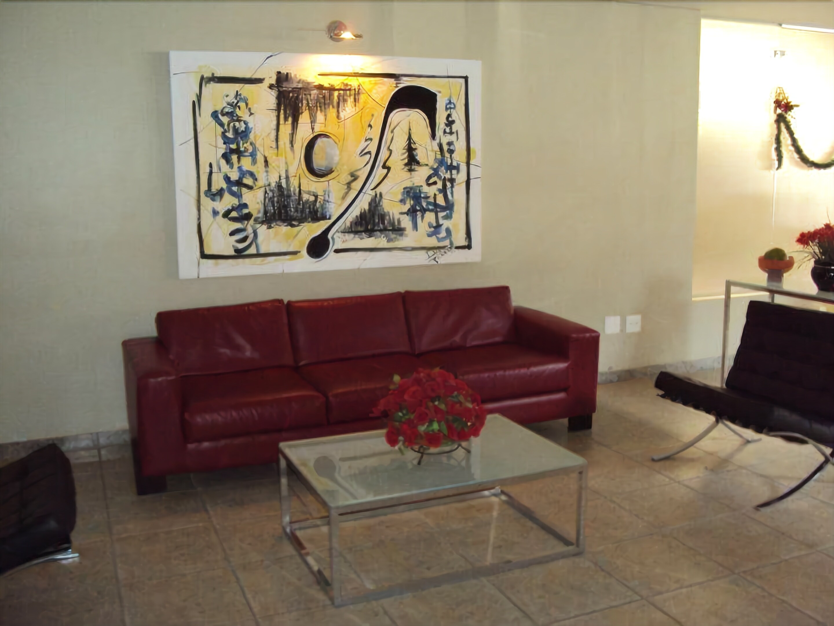 A living room with a red leather sofa, a glass coffee table with a red flower vase, and a large abstract painting on the wall.