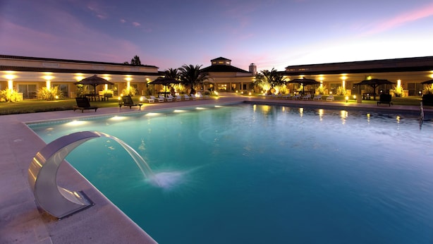 A swimming pool with a water feature, surrounded by lounge chairs and umbrellas, under a building with multiple levels and a flat roof.