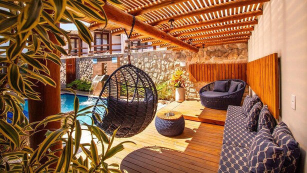 Un'area esterna in legno con una sedia a dondolo, un pouf rotondo e un divano con cuscini decorati.