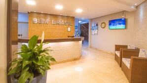 Un'area reception con una scrivania in legno con la scritta "BEM BRASIL", una pianta in vaso, sedie in vimini e un televisore a schermo piatto appeso al muro.