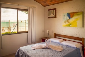 A hotel room with a bed, a window with a view of the beach, a painting on the wall, and an air conditioning unit. - Village Paraíso (Florianópolis)