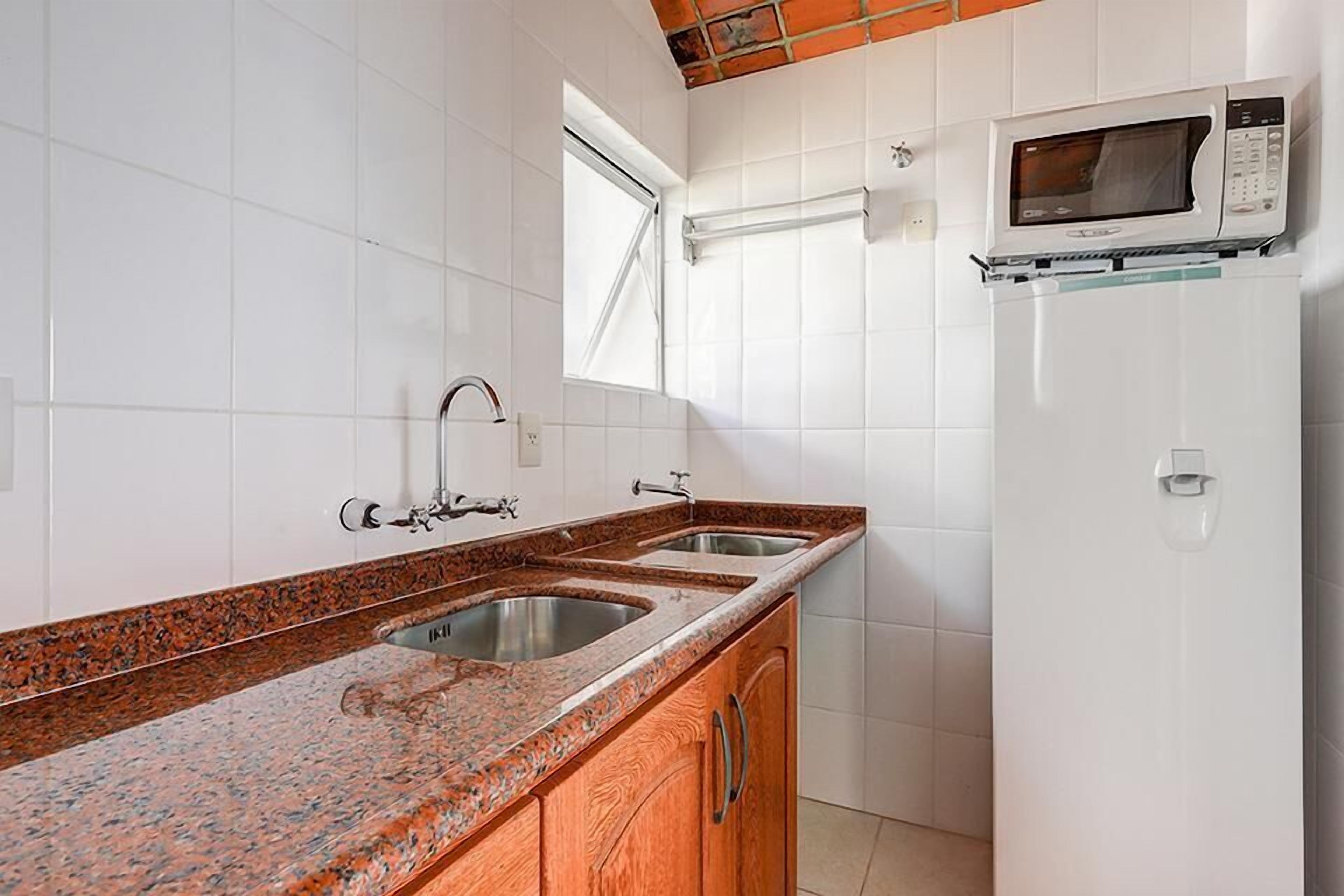 A kitchen with white tiled walls, granite countertops, double sinks, a microwave, and a refrigerator.